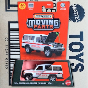 Op de afbeelding: Een Matchbox Moving Parts 2024 Toyota Land Cruiser 70 Series ambulance speelgoedvoertuig in wit met rode accenten. De verpakking bevat een oranje kaart met een afbeelding van het voertuig en een doorzichtige plastic blisterverpakking.