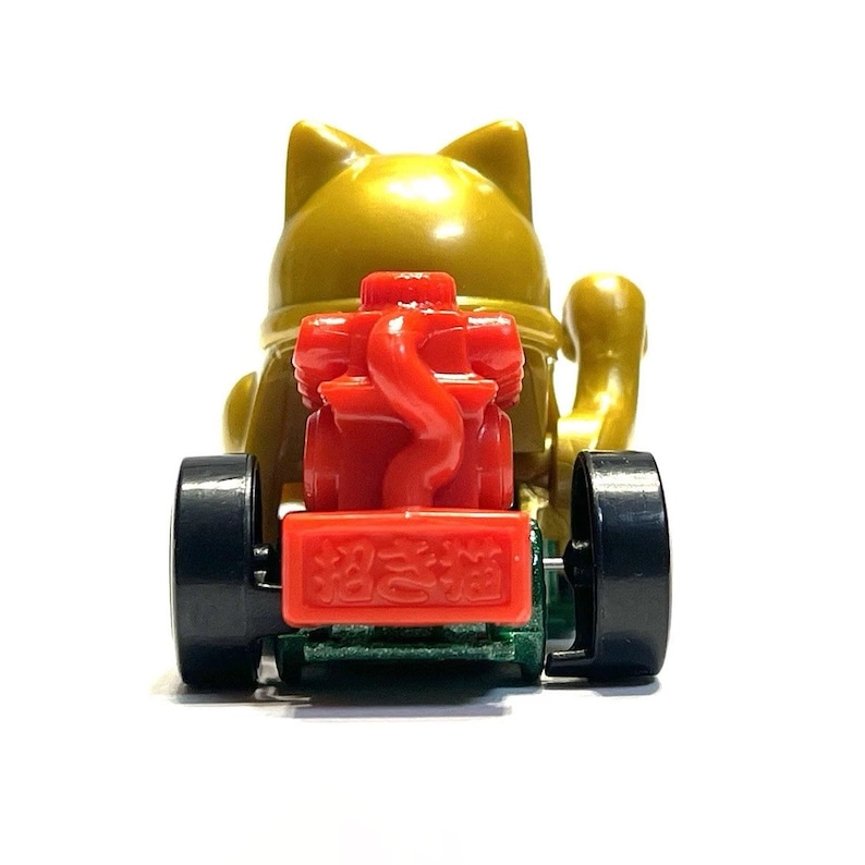 May include: A gold-colored Maneki-neko cat figurine on wheels, with a red engine and black wheels. The figurine has a smiling face and is holding up one paw. The text on the front of the engine is in Japanese.