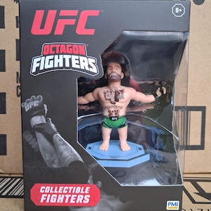Op de afbeelding: Een UFC Octagon Fighters verzamelobject van een mannelijke vechter in een doorzichtige plastic doos. De figuur heeft een baard, tatoeages en een groene korte broek. De doos heeft het UFC-logo en de woorden "Collectable Fighters".