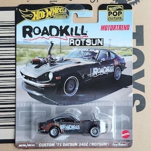May include: A black 1971 Datsun 240Z die-cast toy car with "Roadkill" and "RotSun" graphics. The car is part of the Hot Wheels Premium line and features Real Riders wheels.