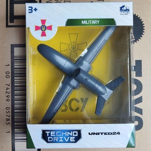 May include: A grey plastic toy aeroplane in a clear plastic package. The package has the text "MILITARY" and "TECHNO DRIVE". The toy is a dark grey colour and has wings and a tail. The package also has a Ukrainian coat of arms.
