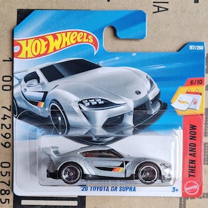 May include: A silver Hot Wheels die-cast toy car of a '20 Toyota GR Supra. The car has black wheels and a black and multi-coloured stripe on the side. The packaging is blue and red, with the text "Hot Wheels" and "Then and Now".