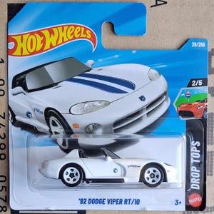 Op de afbeelding: Een witte '92 Dodge Viper RT/10 modelauto met zwarte wielen. De auto heeft blauwe racestrepen en is verpakt in een Hot Wheels blisterverpakking. De verpakking bevat de tekst "DROP TOPS".