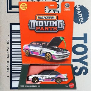 Op de afbeelding: Een Matchbox Moving Parts model van een 1993 Subaru Legacy RS. De auto is wit met roze en blauwe accenten, gouden wielen en de motorkap is open. De verpakking is oranje met het Matchbox logo en de tekst "NEW NOUVEAU 2020".