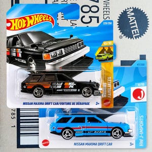 Könnte beinhalten: Zwei Hot Wheels Nissan Maxima Drift Car Modellautos in Originalverpackung. Das obere Fahrzeug ist schwarz mit orangefarbenen und weißen Rennaufklebern, das untere Fahrzeug ist blau mit roten und weißen Streifen. Beide haben detaillierte Räder.