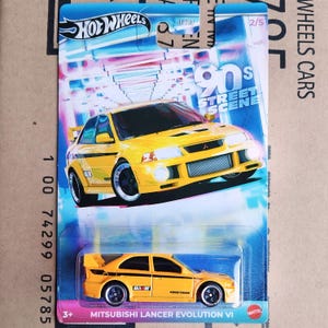 May include: A yellow 1990s Mitsubishi Lancer Evolution VI Hot Wheels die-cast car with black wheels and "Rally Art" and "Toyot Tires" decals on the side. The car is on a blue and pink background with the text "90s Street Scene" and "2/5".