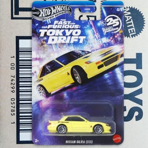 May include: A yellow Nissan Silvia (S13) die-cast model car from the Hot Wheels Silver Series. The packaging features the text "The Fast and the Furious: Tokyo Drift" and a cityscape background. The car is a 1:64 scale model.