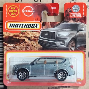 May include: A silver Matchbox die-cast Infiniti QX80 toy SUV in its original orange and red blister pack packaging. The packaging features the Matchbox logo, a Nissan brand mark, and text including New Nouveau 2024 and Designed for a Lifetime.
