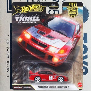 Op de afbeelding: Een rode Hot Wheels Mitsubishi Lancer Evolution VI gegoten auto. De verpakking bevat de tekst "THRILL CLIMBERS" en "10 Car Culture". De auto heeft witte wielen en het nummer 1 aan de zijkant.