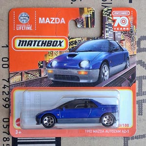 Op de afbeelding: Een blauwe 1992 Mazda Autozam AZ-1 gegoten speelgoedauto in de originele oranje en witte Matchbox-verpakking. De kaart bevat een afbeelding van de auto, het Matchbox 70 Years-logo en de tekst Mazda, Matchbox en 1992 Mazda Autozam AZ-1.