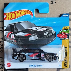 May include: A Hot Wheels Audi 90 Quattro toy car. The black car features red, white, and grey details, with the number 25 visible. The packaging includes the Hot Wheels logo and the text "PEAK PURSUIT".