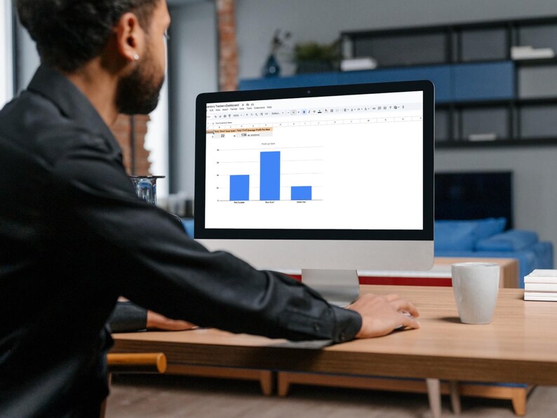 May include: A person working on a desktop computer displaying a bar graph with blue bars. The computer is on a wooden desk, with a white mug and books nearby. The person is wearing a black shirt.