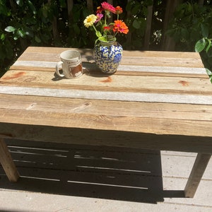 May include: A rustic wooden coffee table with a whitewashed finish. The table has a simple design with four legs and a rectangular top. The top is made of reclaimed wood planks with white painted stripes. A white ceramic mug with a brown bull design and a blue and white ceramic vase with pink and orange flowers are on the table.