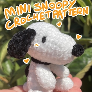 May include: A handmade, mini crochet Snoopy doll, primarily white with black accents. The doll features black ears, a black nose, and a black collar. The text "MINI SNOOPY CROCHET PATTERN" is displayed in orange. The doll is held in a person's hand.