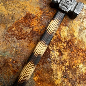 May include: A black-handled hammer with a skull design on the head. The hammer is resting on a rusty metal surface.