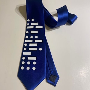 Könnte beinhalten: Eine königsblaue Krawatte mit weißem geometrischen Muster. Die Krawatte besteht aus einem glänzenden Stoff und weist ein sich wiederholendes Design aus weißen Punkten und Rechtecken auf. Die Krawatte liegt auf einer hellen Oberfläche.