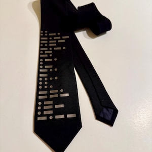 May include: A black tie with a geometric pattern in a lighter shade of gray. The tie is made of a smooth fabric and has a classic pointed end. The design features a series of dots and dashes.
