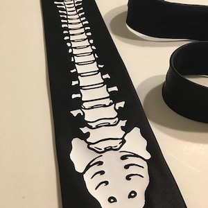 May include: A black tie with a white outline of a human spine. The spine is depicted in anatomical detail, with each vertebra and the rib cage clearly visible.