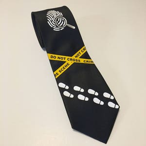 May include: Black tie with a white fingerprint and magnifying glass design. Yellow and black caution tape with the text "Do Not Cross Crime Scene". White footprints are printed on the tie.