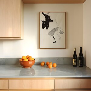 May include: A framed black and white illustration of a couple dancing. The man is holding the woman's hand and they are both jumping in the air. The illustration is hanging on a white wall above a kitchen counter with a bowl of oranges and two bottles of wine.