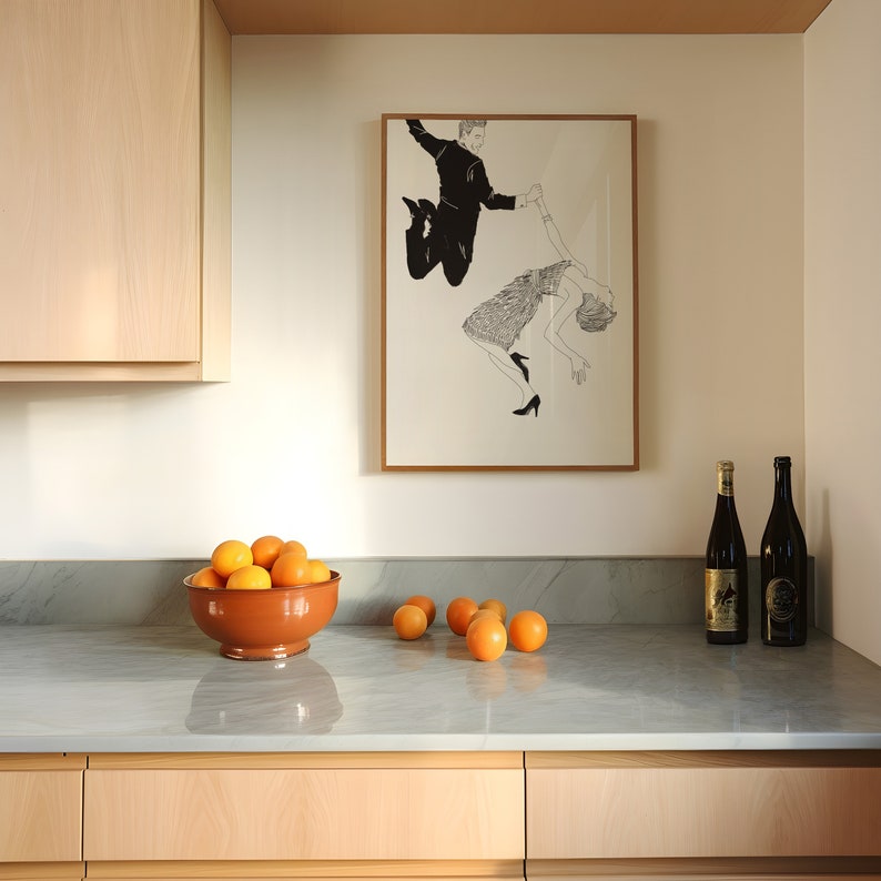 May include: A framed black and white illustration of a couple dancing. The man is holding the woman's hand and they are both jumping in the air. The illustration is hanging on a white wall above a kitchen counter with a bowl of oranges and two bottles of wine.