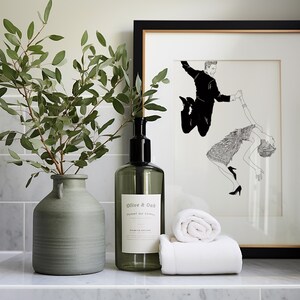 May include: A white bathroom counter with a green vase filled with eucalyptus branches, a clear glass bottle with a black pump labeled "Olive & Oak", and two white towels. A framed black and white illustration of a couple dancing hangs on the wall behind the counter.