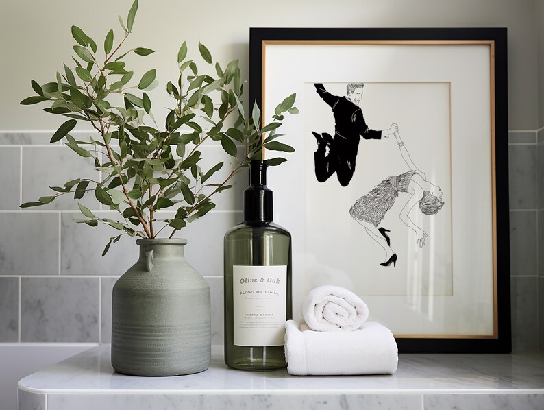 May include: A white bathroom counter with a green vase filled with eucalyptus branches, a clear glass bottle with a black pump labeled "Olive & Oak", and two white towels. A framed black and white illustration of a couple dancing hangs on the wall behind the counter.