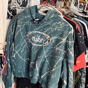 Puede incluir: Una sudadera con capucha Nike de color verde azulado con un dise&ntilde;o de salpicaduras blancas y un logotipo floral de Nike. La sudadera corta est&aacute; colgada en una percha con otras prendas. La sudadera tiene una capucha con cord&oacute;n y una peque&ntilde;a etiqueta Nike.