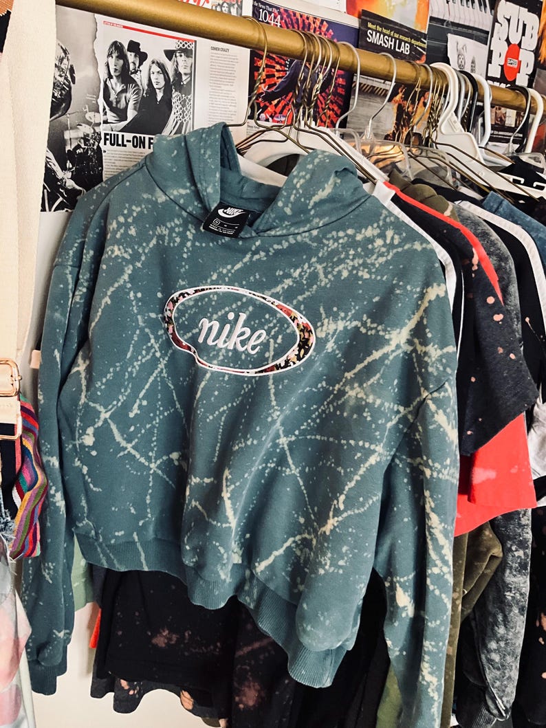 Puede incluir: Una sudadera con capucha Nike de color verde azulado con un dise&ntilde;o de salpicaduras blancas y un logotipo floral de Nike. La sudadera corta est&aacute; colgada en una percha con otras prendas. La sudadera tiene una capucha con cord&oacute;n y una peque&ntilde;a etiqueta Nike.
