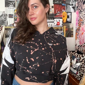 May include: Black cropped hoodie with white accents and a splatter design. The hoodie features the Adidas logo and three stripes on the sleeves. The person is wearing blue jeans. Posters of musicians are in the background.