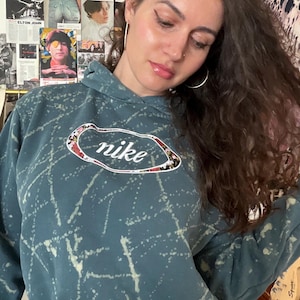 Puede incluir: Sudadera con capucha de color turquesa con dise&ntilde;o de salpicaduras blancas y el logotipo de Nike. El logotipo es un &oacute;valo blanco con el texto Nike y un estampado floral. La sudadera tiene capucha con cord&oacute;n y pu&ntilde;os acanalados.