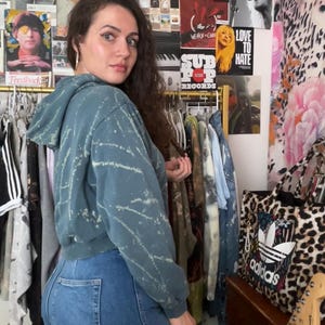 Puede incluir: Una sudadera con capucha corta de color verde azulado con dise&ntilde;o de salpicaduras blancas y vaqueros azules. Detr&aacute;s de la persona hay un perchero con carteles y un bolso con estampado de leopardo. El bolso tiene el logotipo de Adidas.