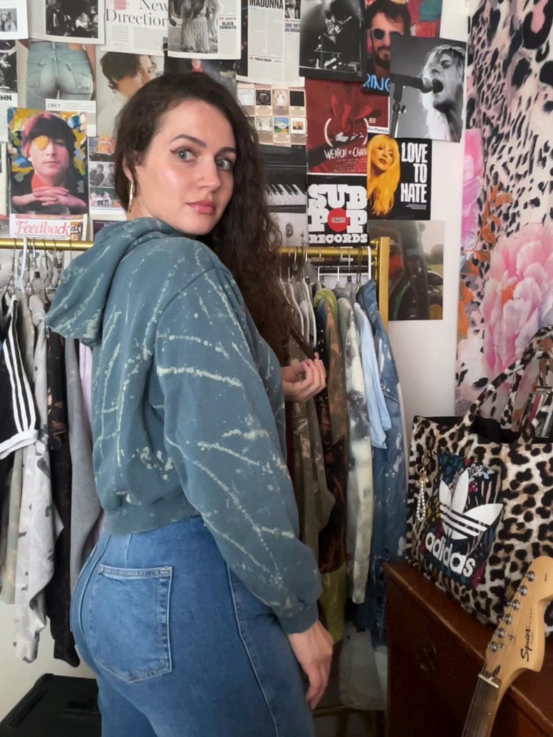 Puede incluir: Una sudadera con capucha corta de color verde azulado con dise&ntilde;o de salpicaduras blancas y vaqueros azules. Detr&aacute;s de la persona hay un perchero con carteles y un bolso con estampado de leopardo. El bolso tiene el logotipo de Adidas.