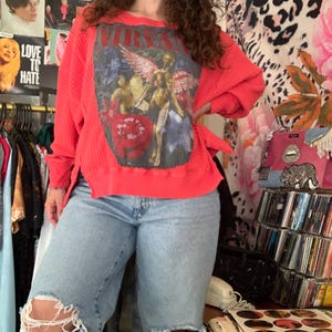 May include: A bright red long-sleeve waffle-knit sweater featuring a large graphic print on the front with the text NIRVANA, a winged figure, and floral elements, paired with light-wash blue denim jeans that have distressed knee holes.