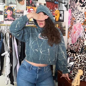 Puede incluir: Una sudadera corta de color verde azulado con dise&ntilde;o de salpicaduras blancas y un logotipo de Nike. La persona lleva vaqueros azules. El fondo presenta carteles y un bolso tote con estampado de leopardo.