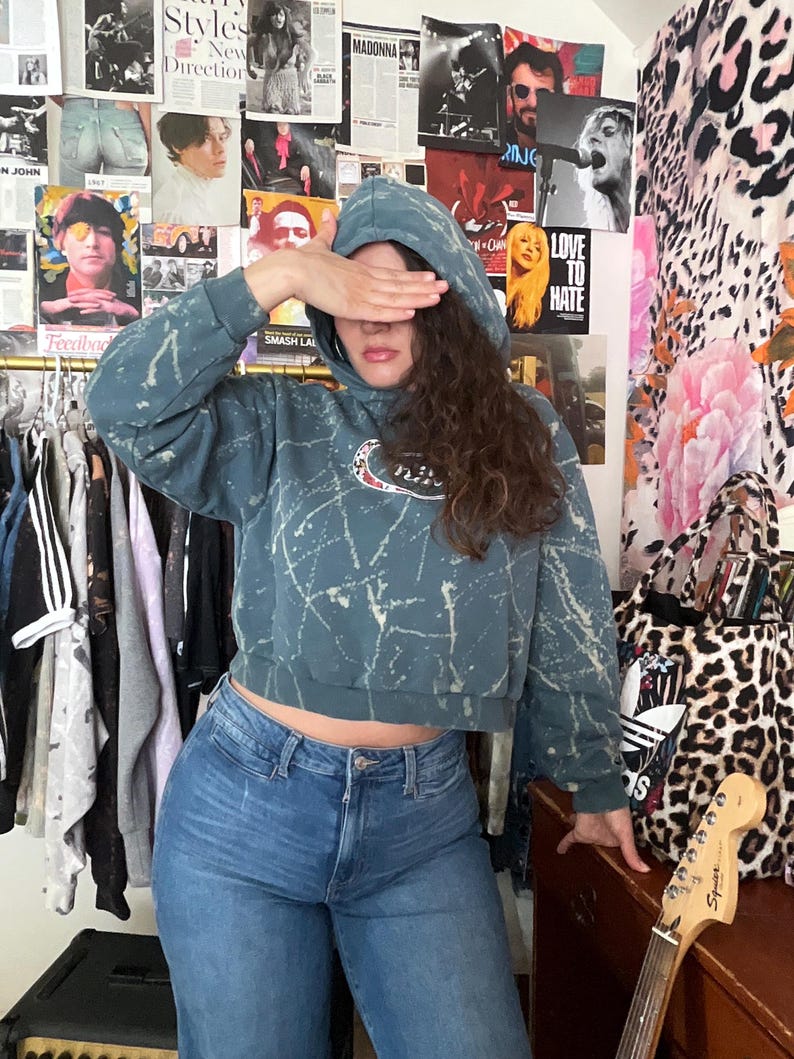 Puede incluir: Una sudadera corta de color verde azulado con dise&ntilde;o de salpicaduras blancas y un logotipo de Nike. La persona lleva vaqueros azules. El fondo presenta carteles y un bolso tote con estampado de leopardo.