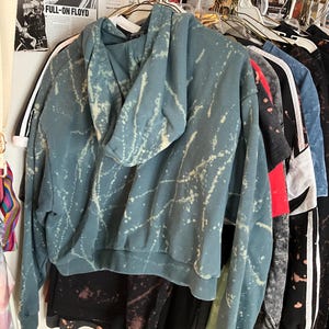 Puede incluir: Una sudadera con capucha de color verde azulado con un dise&ntilde;o de pintura salpicada blanca cuelga de una percha. La sudadera con capucha tiene una capucha con cord&oacute;n y pu&ntilde;os y dobladillo acanalados. Se ven otras prendas, incluidas camisas y sudaderas con capucha.