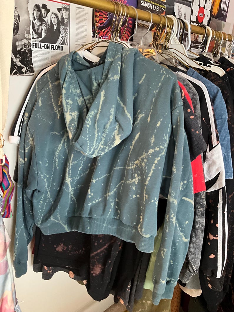 Puede incluir: Una sudadera con capucha de color verde azulado con un dise&ntilde;o de pintura salpicada blanca cuelga de una percha. La sudadera con capucha tiene una capucha con cord&oacute;n y pu&ntilde;os y dobladillo acanalados. Se ven otras prendas, incluidas camisas y sudaderas con capucha.