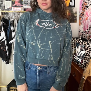 Puede incluir: Una sudadera con capucha corta de color verde azulado con efecto de pintura salpicada blanca y un logotipo de Nike. La sudadera con capucha se combina con unos vaqueros azules. Un bolso tote con estampado de leopardo y una guitarra son visibles en el fondo.