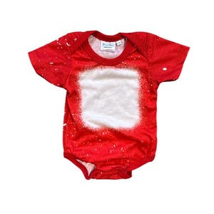 May include: Red baby onesie with a white square in the center, perfect for customization. The onesie has a crew neck and short sleeves. White paint splatter details cover the red fabric.