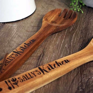 May include: Two wooden kitchen utensils with the text "Sally's Kitchen" engraved on them. The utensils are a slotted spoon and a serving spoon.