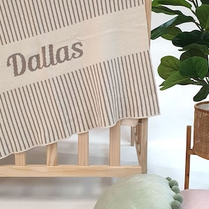 May include: A cream-coloured striped blanket with the name "Dallas" embroidered in brown lettering. The blanket is draped over a wooden cot.