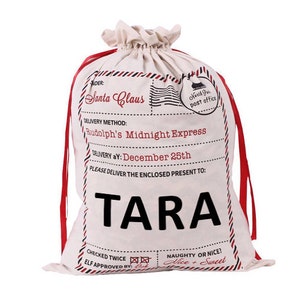 May include: A white canvas Santa sack with red drawstrings. The sack has a printed design that looks like a vintage post office mail bag. The text on the sack reads "Santa Claus", "Rudolph's Midnight Express", "Delivery aY: December 25th", and "Please deliver the enclosed present to: TARA".