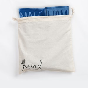May include: A white cotton drawstring bag with the word "thread" printed on the front. The bag is filled with two blue fabric squares with the word "MALJAM" printed on them.