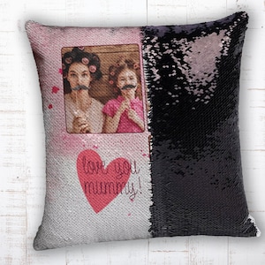 Può includere: Un cuscino rosa e nero con paillettes e una foto di due persone con baffi finti. Il cuscino ha il testo "love you mummy!" in un cuore rosso.