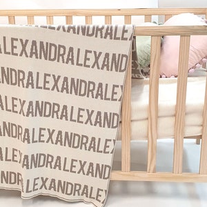 May include: A beige knitted baby blanket with the name "Alexandra" repeated in brown lettering. The blanket is in a wooden crib with a pink pillow.