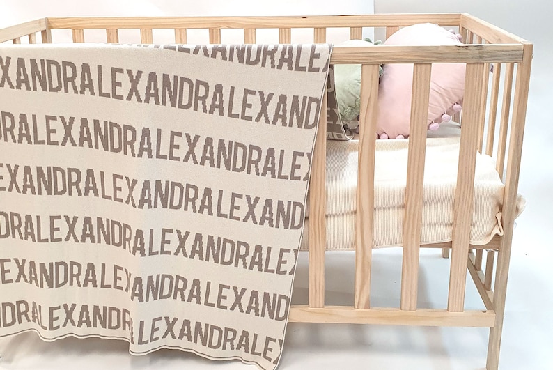 May include: A beige knitted baby blanket with the name "Alexandra" repeated in brown lettering. The blanket is in a wooden crib with a pink pillow.