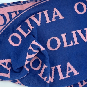 May include: A blue knitted baby blanket with the name "Olivia" repeated in pink lettering.