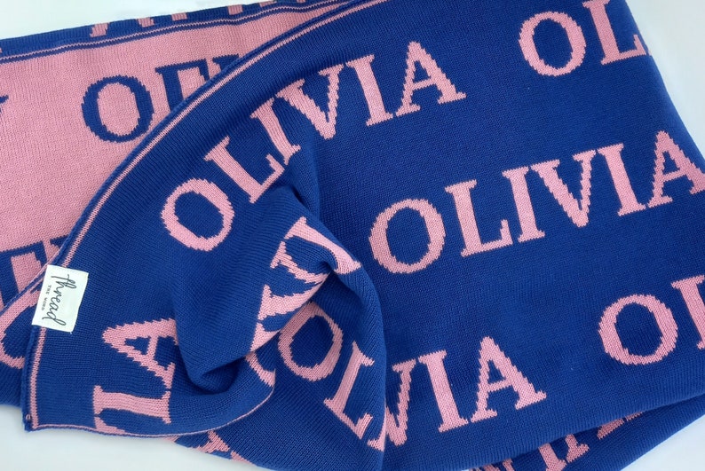 May include: A blue knitted baby blanket with the name "Olivia" repeated in pink lettering.