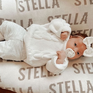May include: A baby wearing a white knitted sweater and white pants lies on a cream-colored blanket with the name "Stella" repeated in brown lettering.